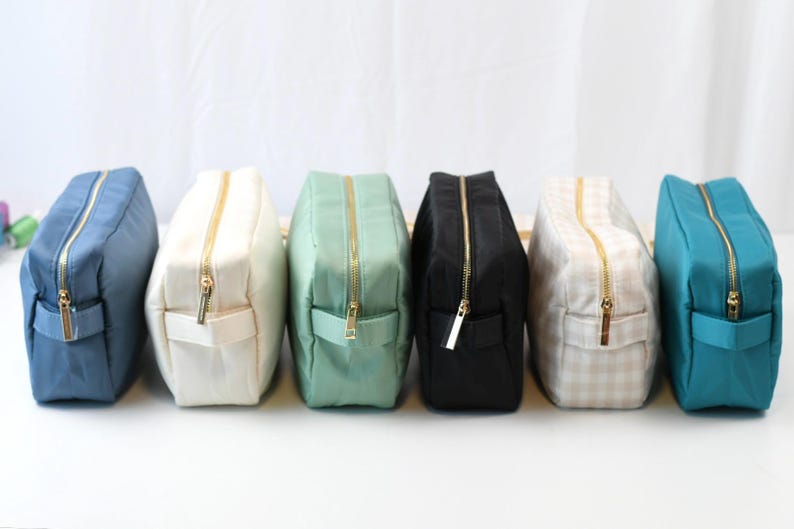 May include: A row of six rectangular zippered pouches in various colors including blue, white, mint green, black, beige gingham, and teal. Each pouch features a gold-toned metal zipper closure and a small fabric side handle for easy carrying.