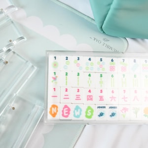 May include: A set of white mahjong tiles with colorful numbers and symbols, including tiles labeled N, E, W, S, and Joker, housed in a clear plastic case. Beside the case are several clear acrylic tile racks on a light blue scalloped mat.