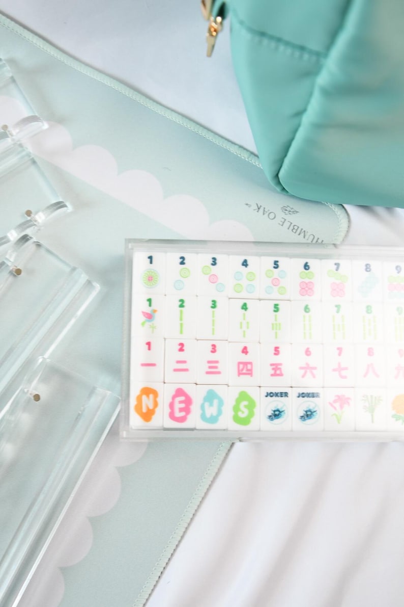 May include: A set of white mahjong tiles with colorful numbers and symbols, including tiles labeled N, E, W, S, and Joker, housed in a clear plastic case. Beside the case are several clear acrylic tile racks on a light blue scalloped mat.
