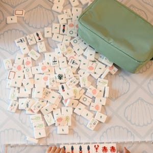 May include: A set of white mahjong tiles featuring various colorful illustrations including numbers, flowers, sea creatures, and sailboats, accompanied by a sage green zippered storage pouch and a clear plastic tile rack.
