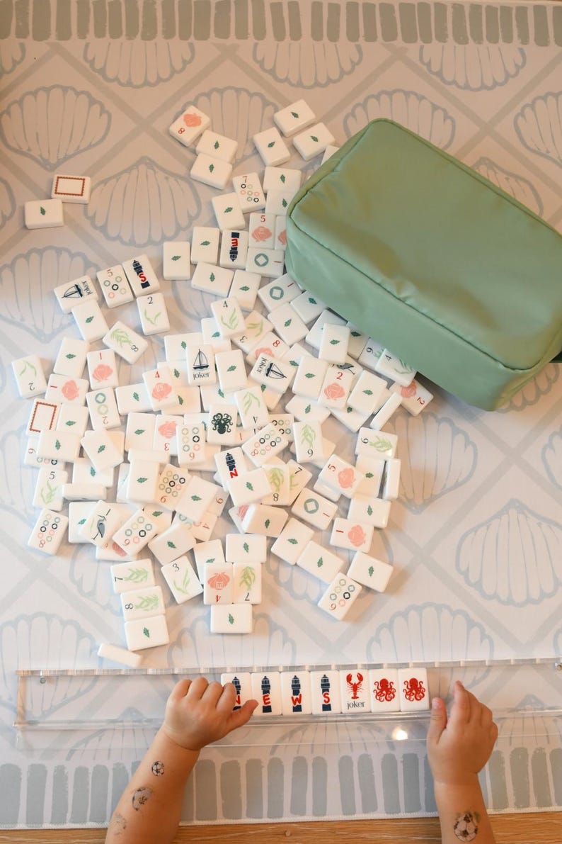 May include: A set of white mahjong tiles featuring various colorful illustrations including numbers, flowers, sea creatures, and sailboats, accompanied by a sage green zippered storage pouch and a clear plastic tile rack.