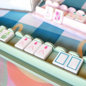 May include: A close-up of a Mahjong set with white tiles featuring pink and green designs. The tiles are arranged in light green trays on a blue and white checkered surface. A clear plastic container holds additional tiles.