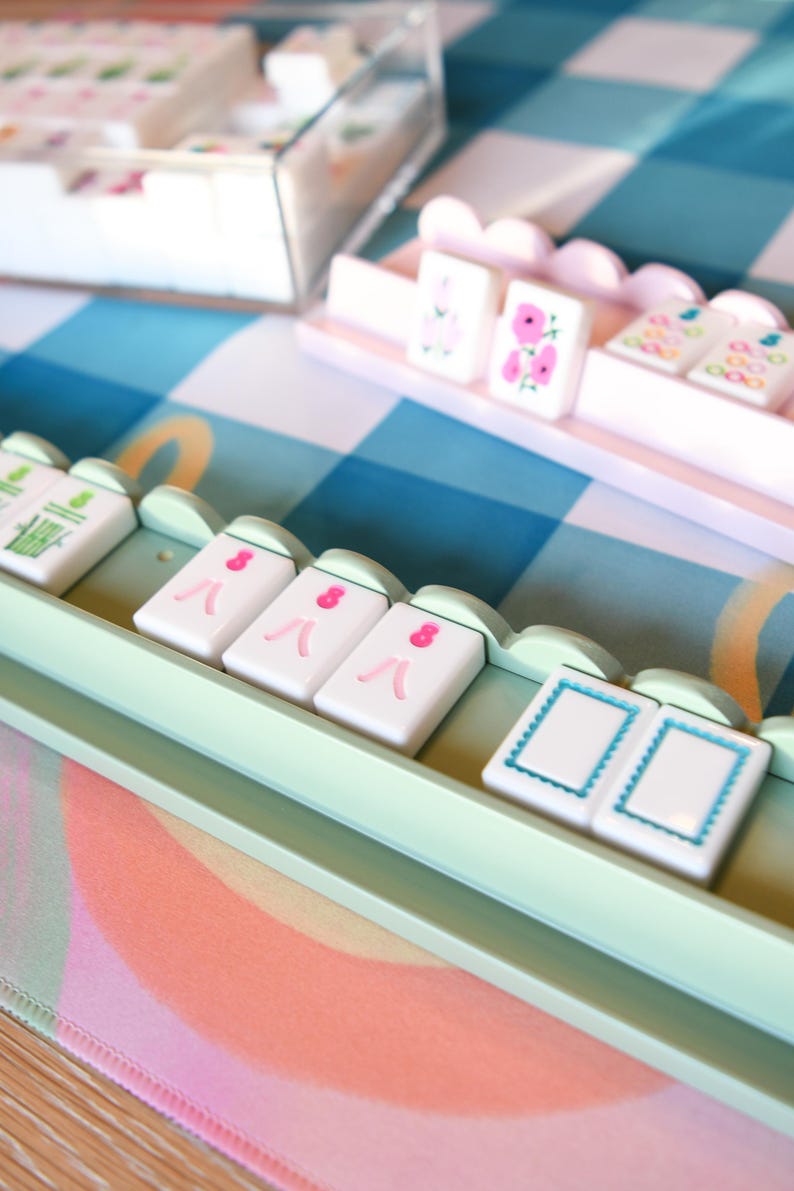 May include: A close-up of a Mahjong set with white tiles featuring pink and green designs. The tiles are arranged in light green trays on a blue and white checkered surface. A clear plastic container holds additional tiles.