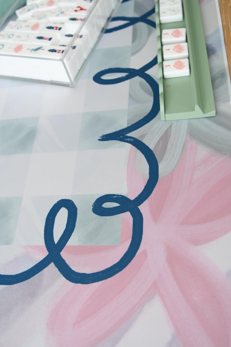 May include: A colorful game mat with a floral design in pink and blue, featuring a navy blue swirl. A clear box and a green tray hold game pieces with illustrations. The background has a checkered pattern.