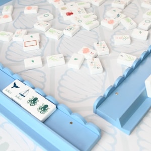 May include: A rummy tile game set featuring white rectangular tiles with various colorful illustrations and numbers, scattered on a light blue and white seashell-patterned board. Two light blue wooden tile racks with scalloped edges are included.