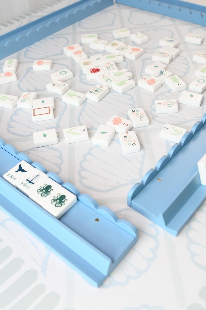May include: A rummy tile game set featuring white rectangular tiles with various colorful illustrations and numbers, scattered on a light blue and white seashell-patterned board. Two light blue wooden tile racks with scalloped edges are included.