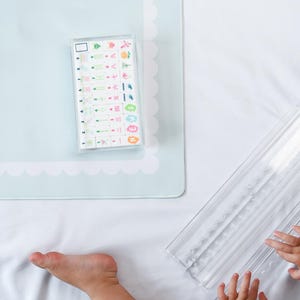 May include: A light blue rectangular mat with a scalloped white border and the text HUMBLE OAK, featuring a clear plastic case containing a set of small white tiles with colorful numbers, symbols, and Japanese characters, plus a clear tile rack.