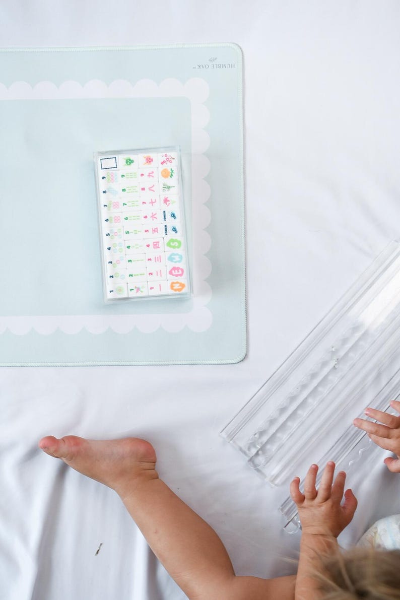 May include: A light blue rectangular mat with a scalloped white border and the text HUMBLE OAK, featuring a clear plastic case containing a set of small white tiles with colorful numbers, symbols, and Japanese characters, plus a clear tile rack.