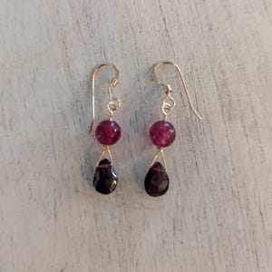 May include: A pair of gold-colored dangle earrings. Each earring features a round, deep red bead above a dark, teardrop-shaped gem. The earrings hang from gold-colored ear wires.
