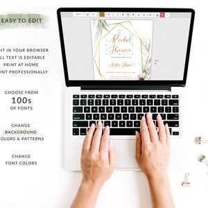 May include: A laptop computer with a screen displaying a customizable bridal shower invitation template. The invitation features a gold geometric frame with green foliage and the text "It's a Bridal Shower" in a gold script font. The text "Honoring Bride-to-Be" is in a smaller font below the main text. The invitation also includes the bride's name, the date, time, and location of the shower. The laptop is on a white surface with paper clips.