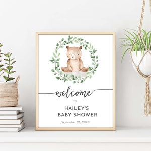 May include: A printable welcome sign for a baby shower with a watercolor illustration of a brown bear sitting in a green leafy wreath. The text reads "Welcome to Hailey's Baby Shower September 23, 2020".