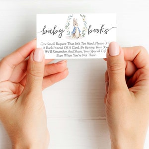 May include: A white card with the text "baby books" in black script. The card features a watercolor illustration of a rabbit wearing a blue jacket and holding a book. The text on the card reads: "One small request that isn't too hard, please bring a book instead of a card. By signing your book, we'll remember and share, your special gift even when you're not there."