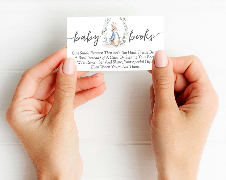 May include: A white card with the text "baby books" in black script. The card features a watercolor illustration of a rabbit wearing a blue jacket and holding a book. The text on the card reads: "One small request that isn't too hard, please bring a book instead of a card. By signing your book, we'll remember and share, your special gift even when you're not there."