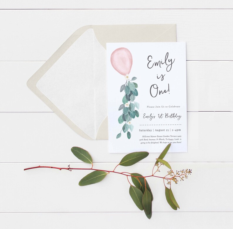 May include: A white invitation card with a pink balloon and green eucalyptus leaves. The text on the card reads "Emily is One!" and includes details for Emily's 1st birthday party.