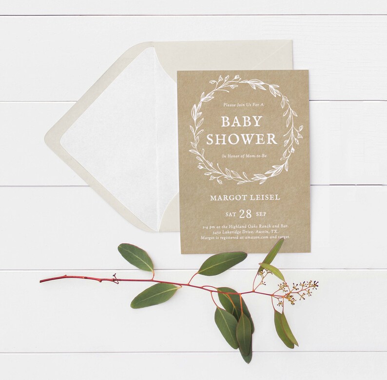 May include: A brown kraft paper baby shower invitation with a white wreath design and the text "Please Join Us For A Baby Shower In Honor of Mom-to-Be Margot Leisel Sat 28 Sep 2-4 pm at the Highland Oaks Ranch and Bar 5415 Lakeridge Drive, Austin, TX Margot is registered at amazon.com and target."