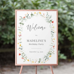 May include: Welcome to Madeline's Birthday Party sign with a floral wreath border in shades of pink, yellow, and green. The sign is on a wooden easel.