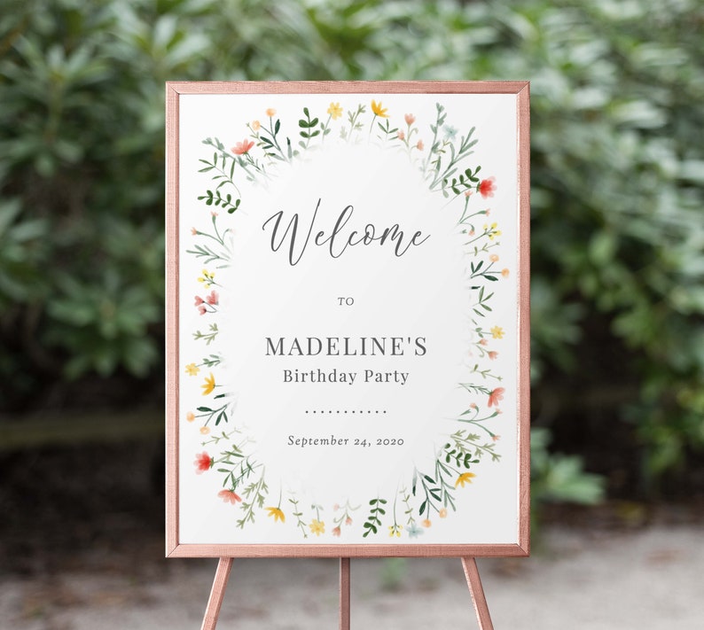 May include: Welcome to Madeline's Birthday Party sign with a floral wreath border in shades of pink, yellow, and green. The sign is on a wooden easel.