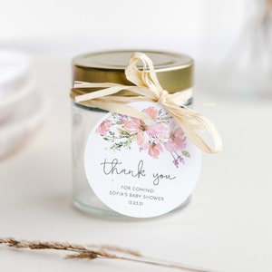 May include: A round, white thank you tag with pink watercolor flowers and the text "thank you for coming! Sofia's Baby Shower 12.23.21" tied to a glass jar with a gold lid and a beige ribbon.