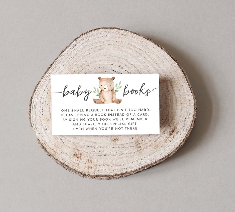 May include: A white card with the text "baby books" in black cursive script. The card features a small illustration of a brown bear with green leaves. The text on the card reads: "One small request that isn't too hard, please bring a book instead of a card. By signing your book we'll remember and share, your special gift, even when you're not there."