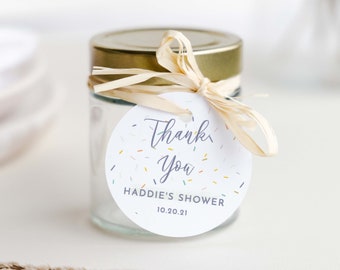 May include: A round white thank you tag with colourful confetti and the text "Thank You" in purple script. The tag also says "Haddie's Shower 10.20.21". The tag is tied to a glass jar with a gold lid and a beige ribbon.
