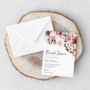 Op de afbeelding: Een witte envelop met een bloemige bruidsdoucheinvitatie. De uitnodiging heeft een aquarelontwerp met roze en bordeauxrode bloemen met groene bladeren. De tekst op de uitnodiging luidt "Bridal Shower" en "Wordt u alstublieft bij ons om de toekomstige bruid Katie Timmons te verwennen om 14:00 uur in de Hillcrest Manor Estate Garden Terrace tegenover Bond Avenue in Wood, TX. Proost! Cadeaulijst: Babylist en Amazon.com"