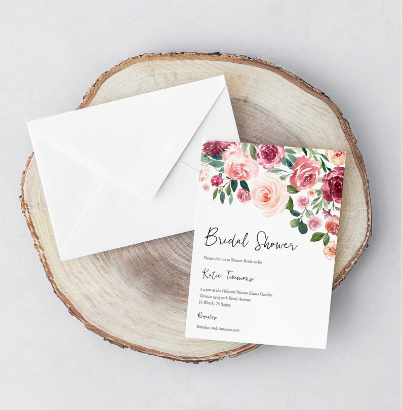 Op de afbeelding: Een witte envelop met een bloemige bruidsdoucheinvitatie. De uitnodiging heeft een aquarelontwerp met roze en bordeauxrode bloemen met groene bladeren. De tekst op de uitnodiging luidt "Bridal Shower" en "Wordt u alstublieft bij ons om de toekomstige bruid Katie Timmons te verwennen om 14:00 uur in de Hillcrest Manor Estate Garden Terrace tegenover Bond Avenue in Wood, TX. Proost! Cadeaulijst: Babylist en Amazon.com"