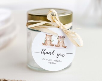 May include: A round white thank you tag with two brown bears wearing floral crowns. The tag is tied to a clear glass jar with a gold lid and a beige ribbon. The tag says "thank you" in cursive and "Olivia's Shower 10.20.20".