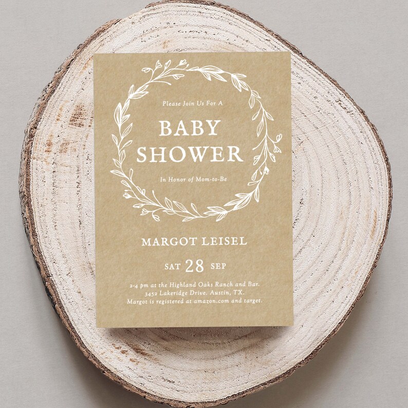 May include: A kraft paper baby shower invitation with a white floral wreath and the text "Please Join Us For A Baby Shower In Honor of Mom-to-Be Margot Leisel Sat 28 Sep 2-4 pm at the Highland Oaks Ranch and Bar. 3452 Lakeridge Drive. Austin, TX. Margot is registered at amazon.com and target."