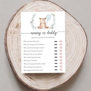 Puede incluir: Una tarjeta de juego para baby shower con el texto "mam&aacute; o pap&aacute;" y una ilustraci&oacute;n en acuarela de un oso de peluche sosteniendo un globo azul. La tarjeta incluye preguntas con iconos de bigote y labios.