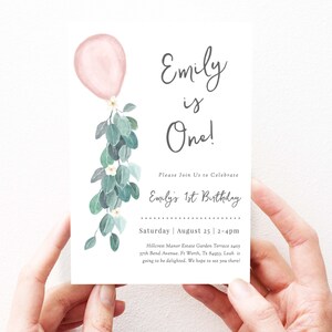 May include: A white invitation card with a pink balloon and green eucalyptus leaves. The text on the card reads "Emily is One!" and includes details for a first birthday party.