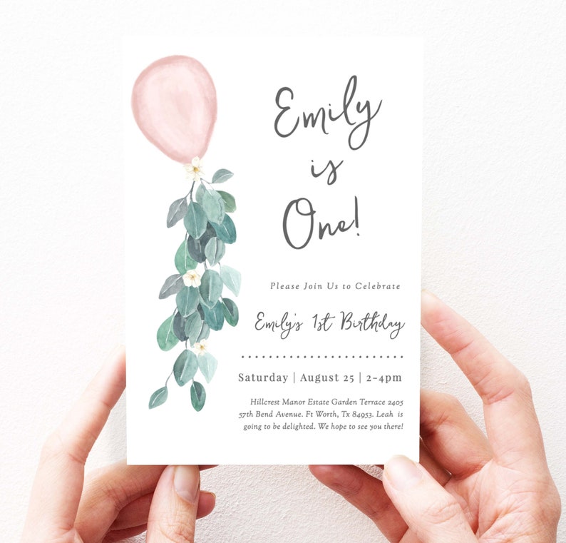 May include: A white invitation card with a pink balloon and green eucalyptus leaves. The text on the card reads "Emily is One!" and includes details for a first birthday party.