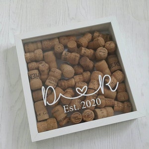 May include: White shadow box with a glass front filled with cork stoppers. The box has the words "Our" and "Est. 2020" written on it.