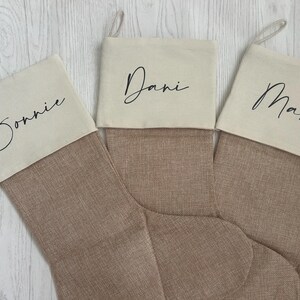 May include: Three Christmas stockings featuring a tan, textured fabric body and a cream-coloured cuff. Each cuff is personalised with black script text reading Bonnie, Dani, and Max. Each stocking includes a small fabric loop for hanging purposes.