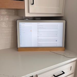 Peut inclure: Une tablette numérique avec un cadre noir et un support en bois clair affiche un menu de paramètres. La tablette est posée sur un comptoir blanc avec un motif marbré, sous des armoires blanches avec des poignées noires. Une étagère en bois au-dessus contient des bouchons.