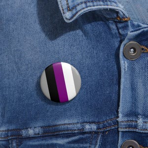 Pode incluir: Um botão redondo com um design listrado preto, roxo, branco e cinza. O botão está em uma jaqueta jeans azul.