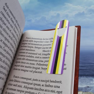 Puede incluir: Un marcapáginas a rayas de color morado, amarillo y negro con una cara sonriente blanca. El marcapáginas está en un libro con texto sobre un tipo de planta.