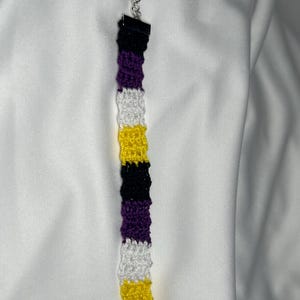 Könnte beinhalten: Ein gehäkeltes Armband mit gelben, weißen, violetten und schwarzen Streifen. Das Armband hat an beiden Enden silberne Verschlüsse.