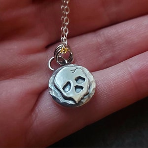 May include: A silver necklace with a small, round pendant featuring a skull design. The pendant has a textured surface and the skull is slightly raised.