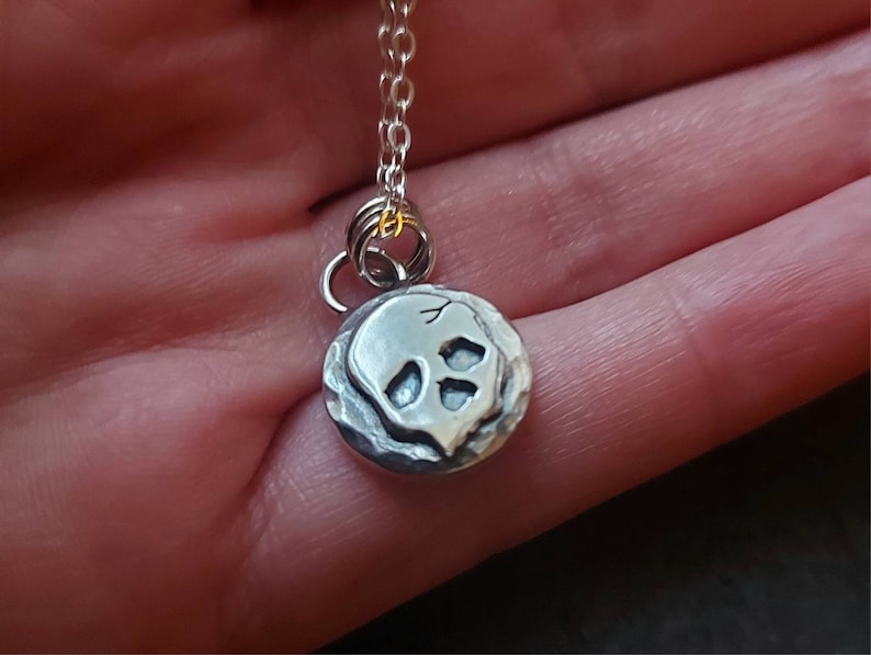May include: A silver necklace with a small, round pendant featuring a skull design. The pendant has a textured surface and the skull is slightly raised.