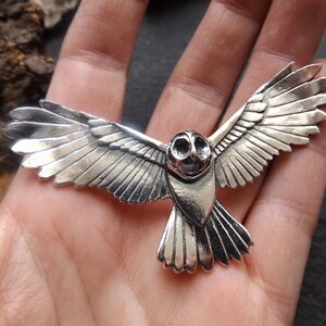 May include: A silver owl pendant with detailed wings spread in flight. The owl's eyes are large and round, and its beak is small and pointed.