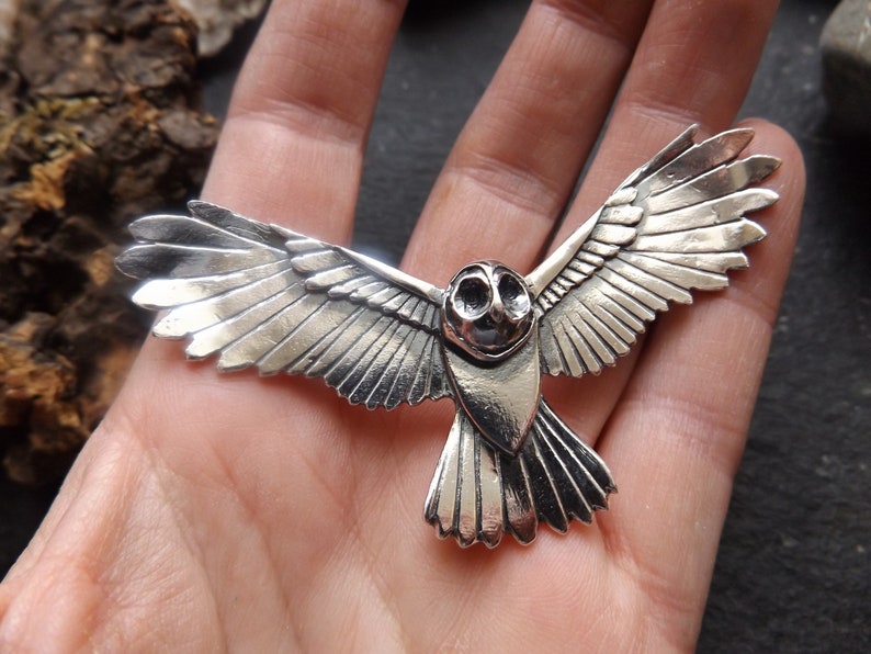 May include: A silver owl pendant with detailed wings spread in flight. The owl's eyes are large and round, and its beak is small and pointed.