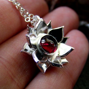 May include: A silver pendant necklace featuring a red gemstone set in a flower-shaped design. The pendant is detailed with intricate carvings and a polished finish.