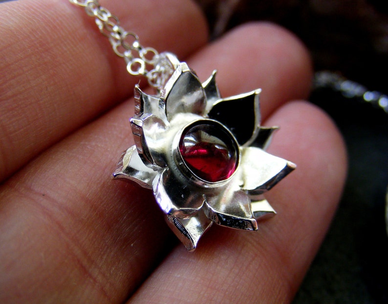 May include: A silver pendant necklace featuring a red gemstone set in a flower-shaped design. The pendant is detailed with intricate carvings and a polished finish.