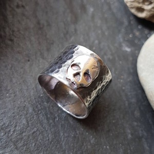 May include: A silver ring with a hammered texture and a skull design. The skull is made of a different metal, possibly gold.