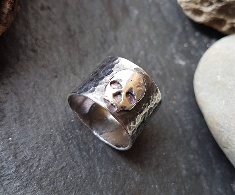 May include: A silver ring with a hammered texture and a skull design. The skull is made of a different metal, possibly gold.