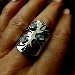 May include: A silver ring with a tree design and three moonstone gems. The ring is worn on a finger.