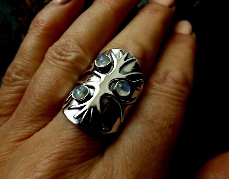 May include: A silver ring with a tree design and three moonstone gems. The ring is worn on a finger.