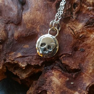 May include: A silver necklace with a small, round pendant featuring a skull design. The pendant is textured and has a slightly hammered look.