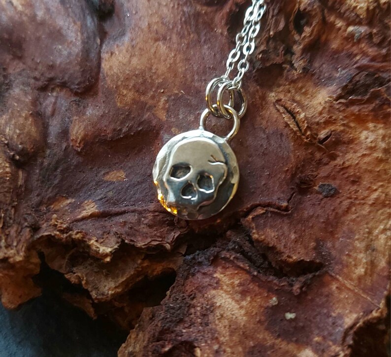 May include: A silver necklace with a small, round pendant featuring a skull design. The pendant is textured and has a slightly hammered look.
