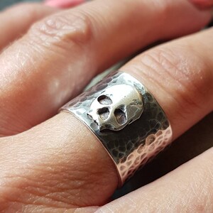May include: A silver ring with a hammered finish and a skull design. The ring is worn on a finger.